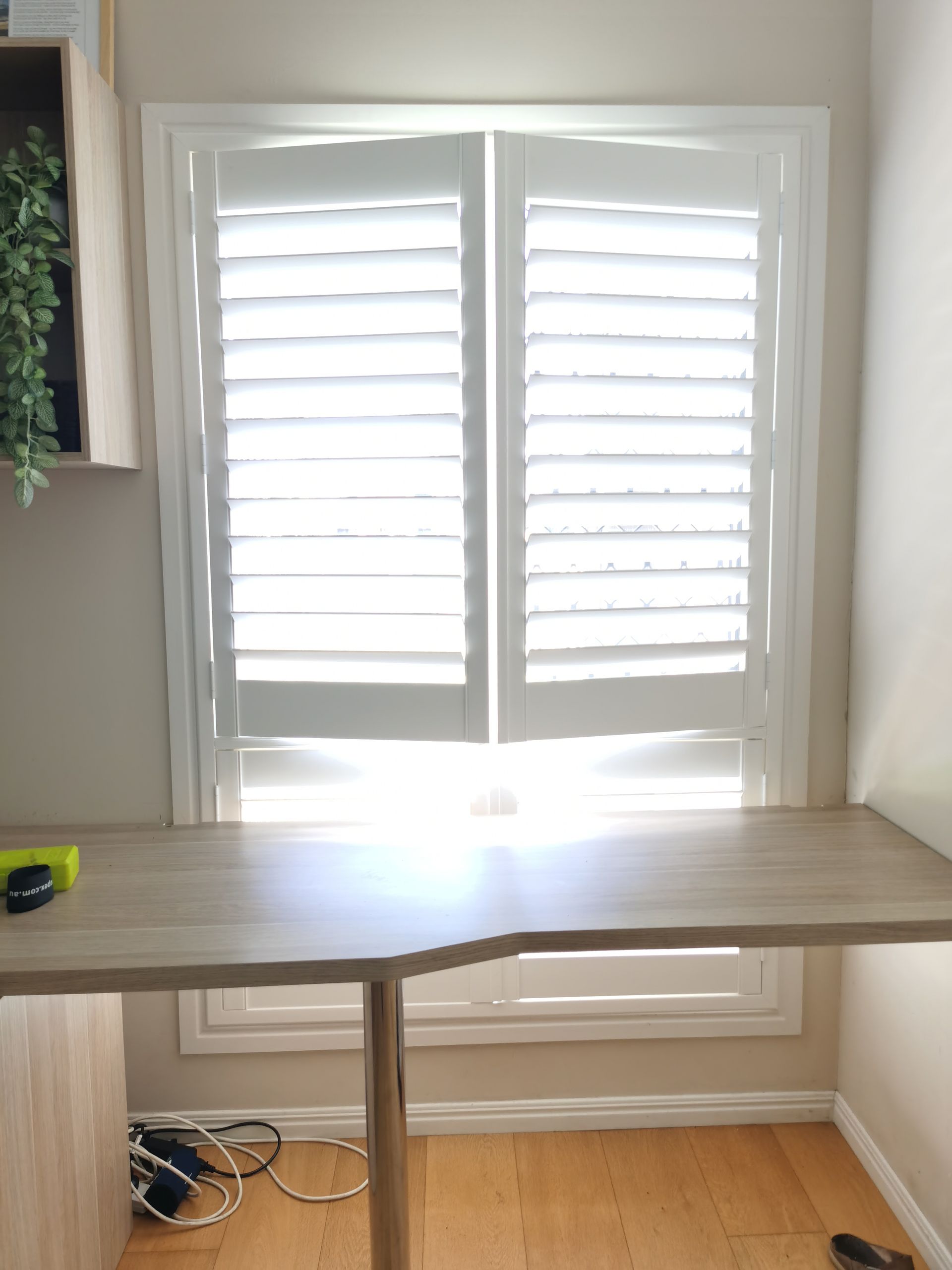 There is a desk in front of a window with white shutters.