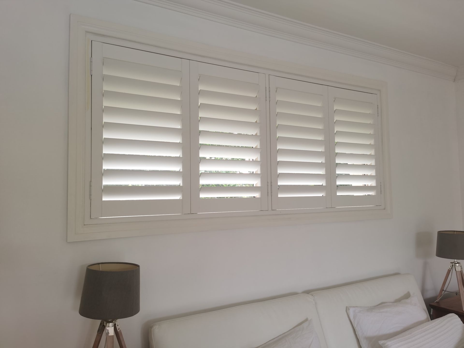 A bedroom with white shutters on the window and a lamp.