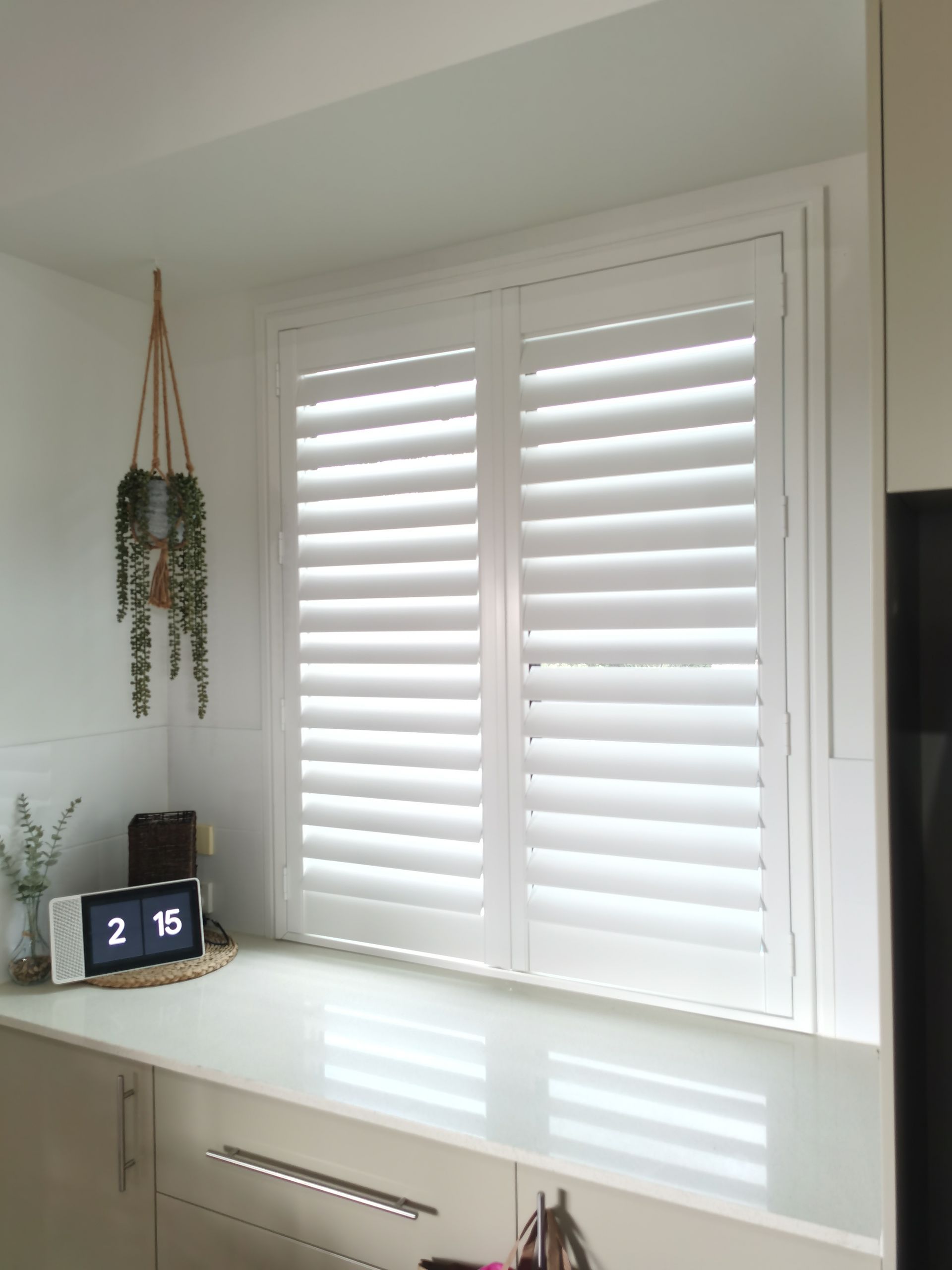 A kitchen window with white shutters and a clock on the counter.