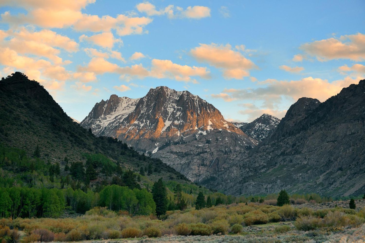 Mountain landscape with snow-dusted peak, forested valley, and cloudy sunset sky