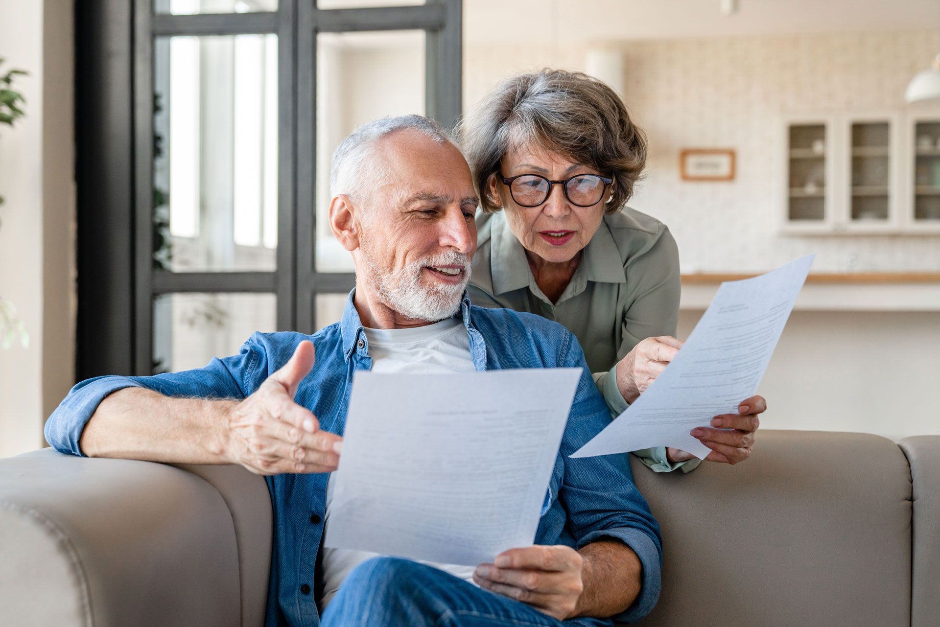 Senior couple reviewing documents together on a couch, indoors. The man points to the paper.