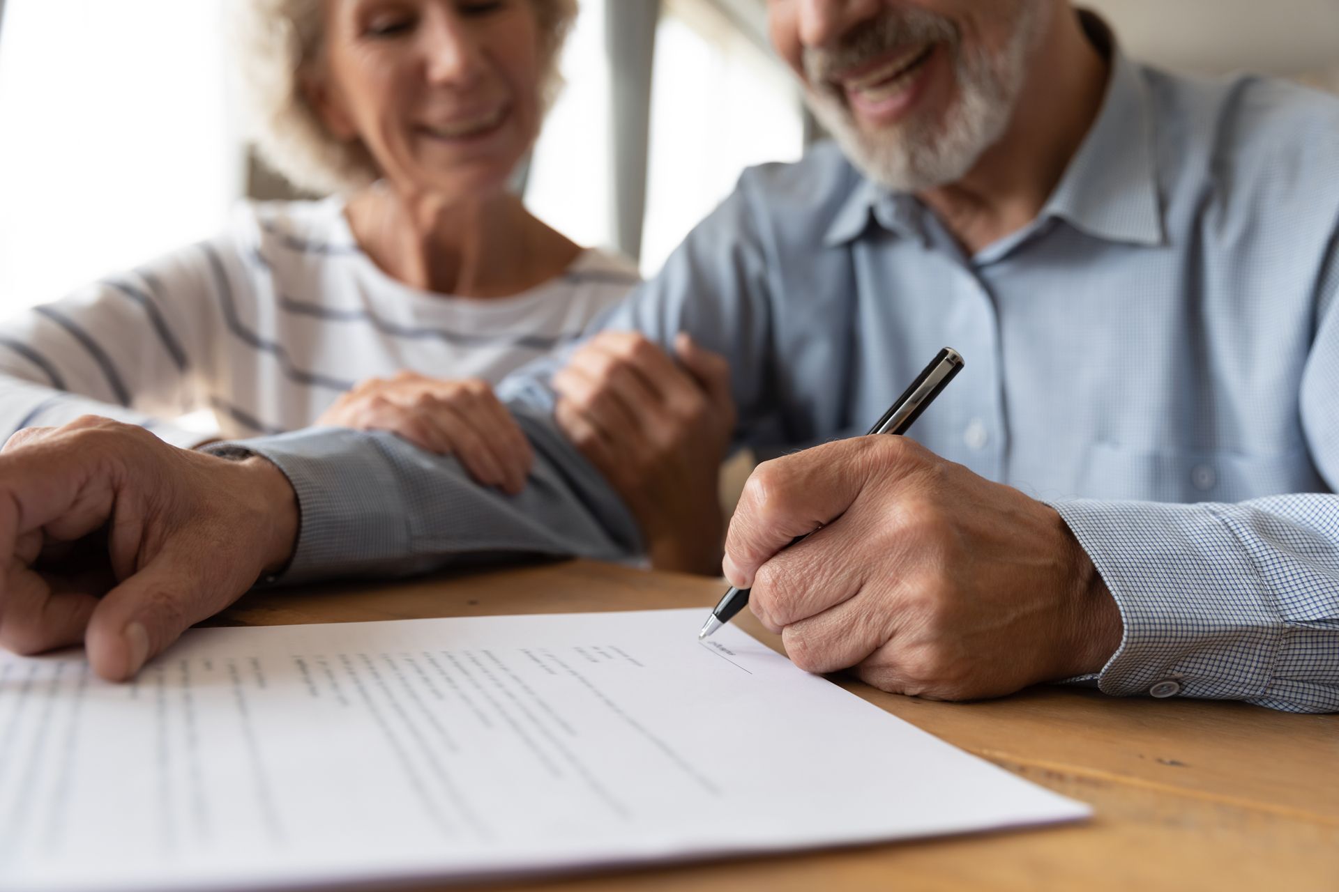 Senior man signing document, woman smiles and touches his shoulder at a table