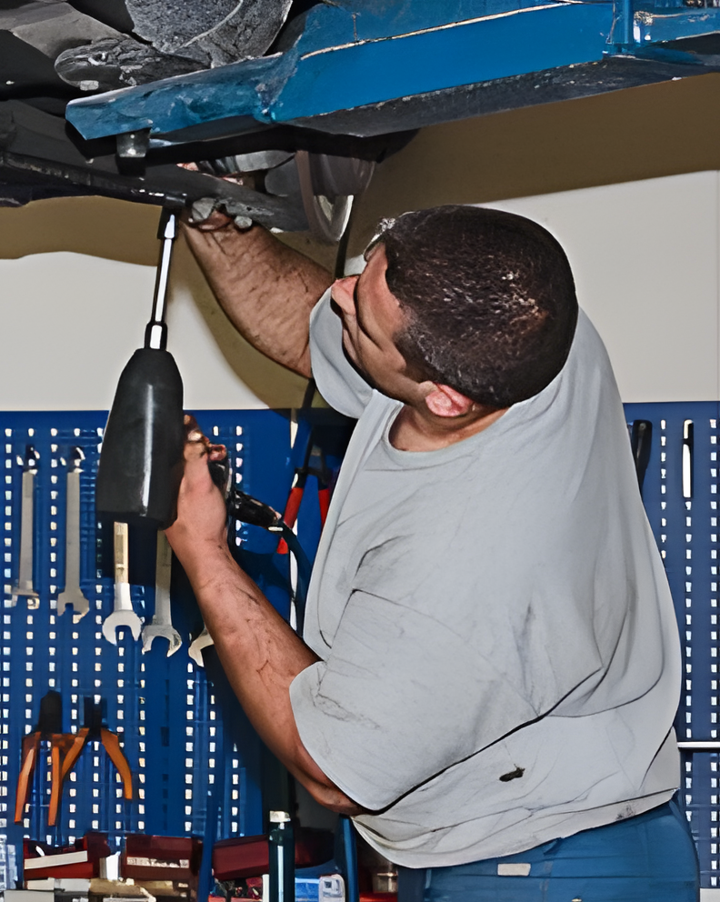 Mechanic using a tool under a car in a garage. Blue wall with tools in background. | Discount Brake & Auto Repair