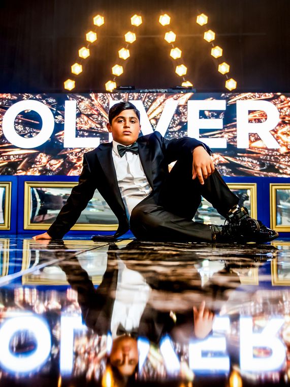 Person posing on stage in front of OLIVER sign, seated on reflective floor with bright lights behind.