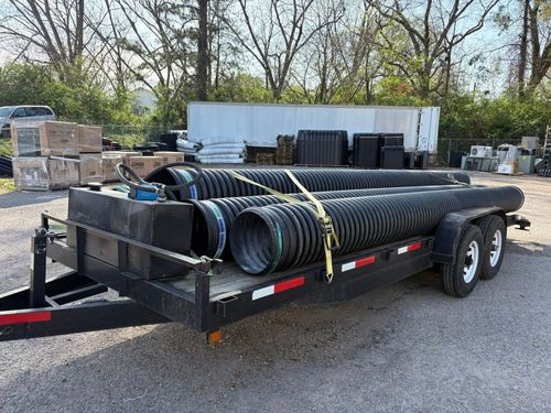 Black utility trailer loaded with large black drainage pipes in an outdoor gravel lot