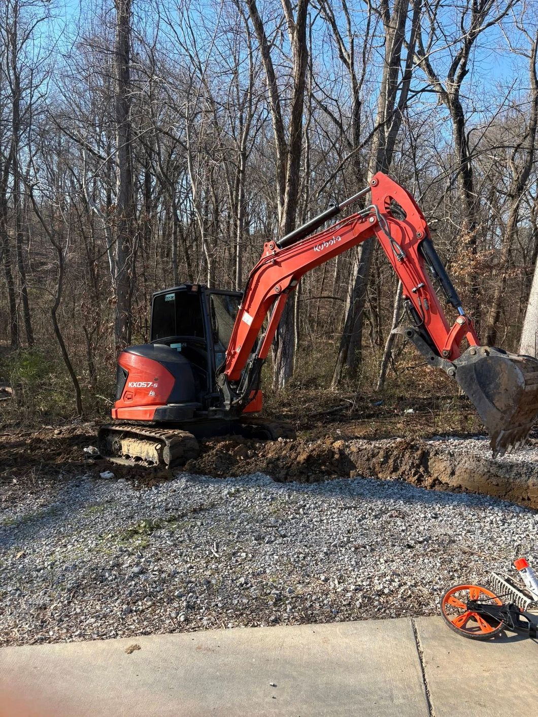 Red excavator working on a gravel area beside a concrete road in a wooded setting