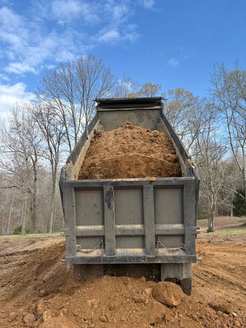 Dump truck unloading a mound of dirt at a wooded outdoor site under a blue sky