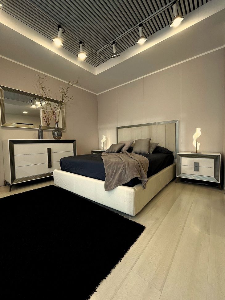 Camera da letto moderna con biancheria da letto grigia, testiera bianca, specchio e vaso di rami su un comodino.
