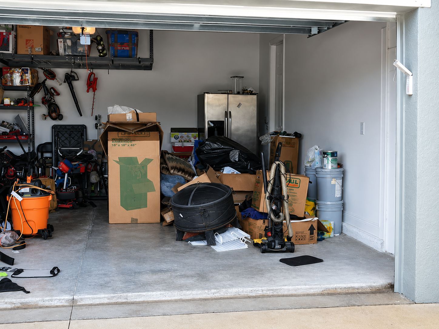 Cluttered garage with tools, boxes, bikes, and stored items on a concrete floor.