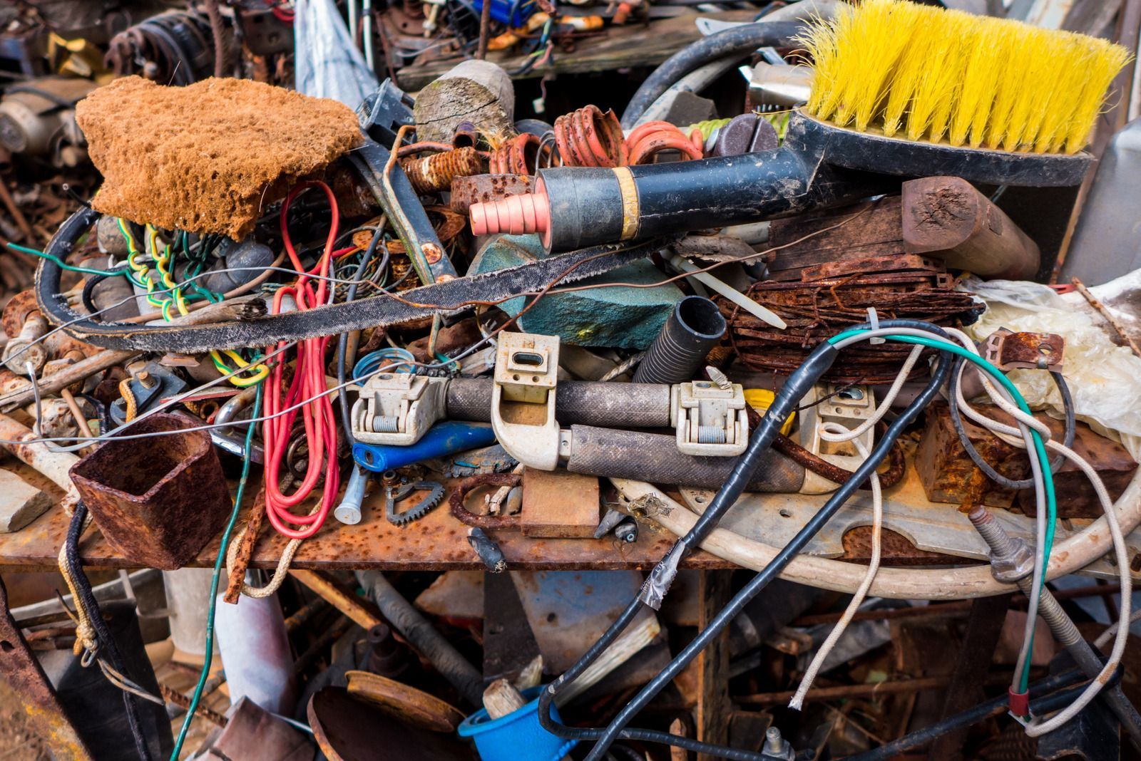 A cluttered workbench filled with assorted scrap metal, rusty tools, wiring, and a yellow-bristled brush.
