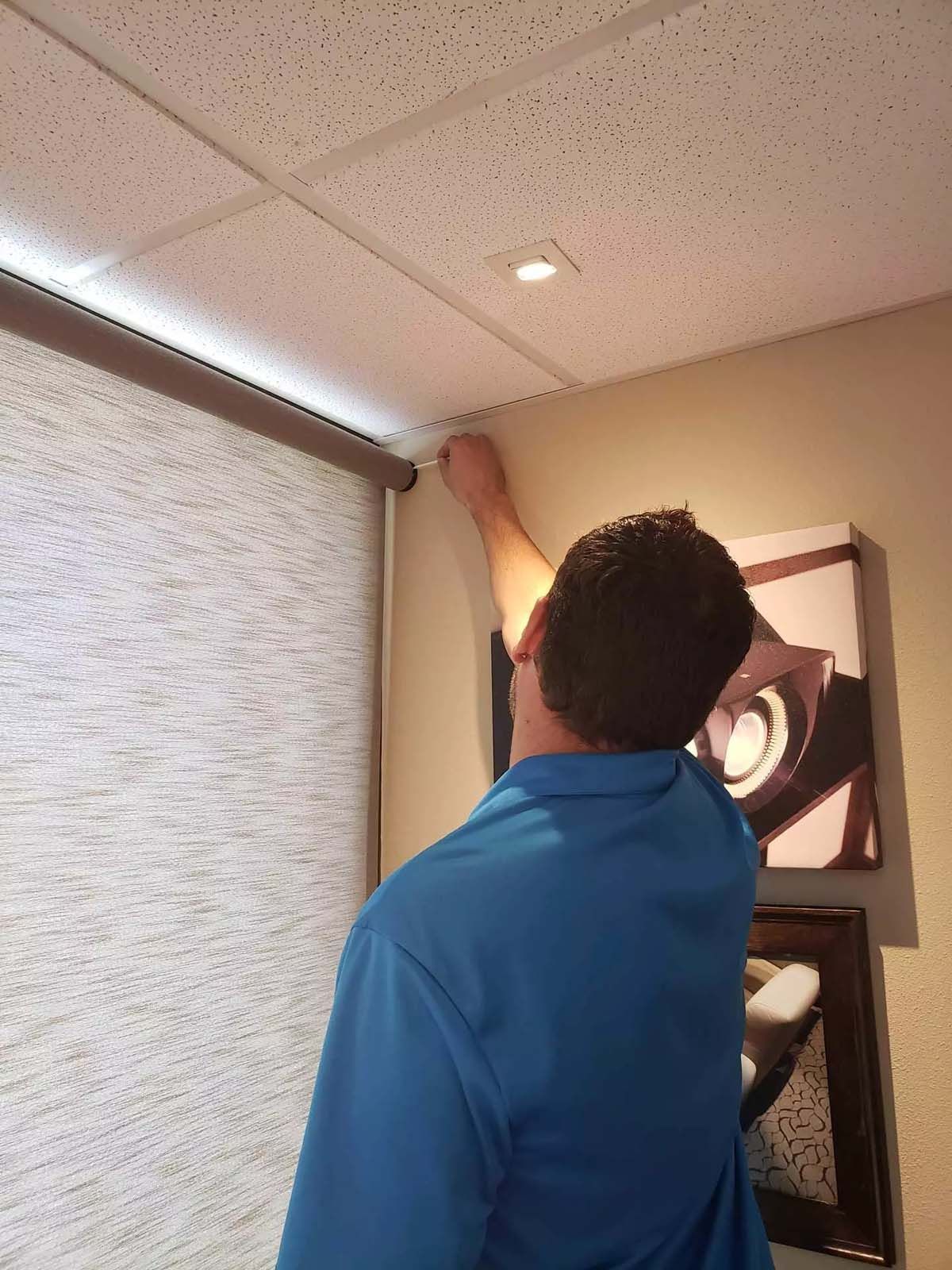A man in a blue shirt is adjusting a blind on a window.
