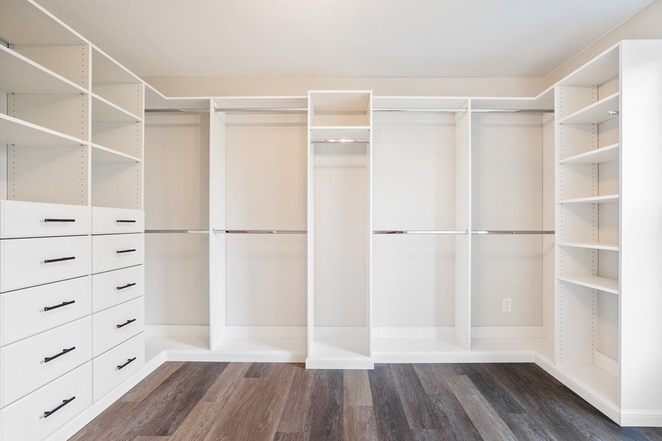 Empty white walk-in closet with shelves, drawers, and hanging rods. Dark wood floor.