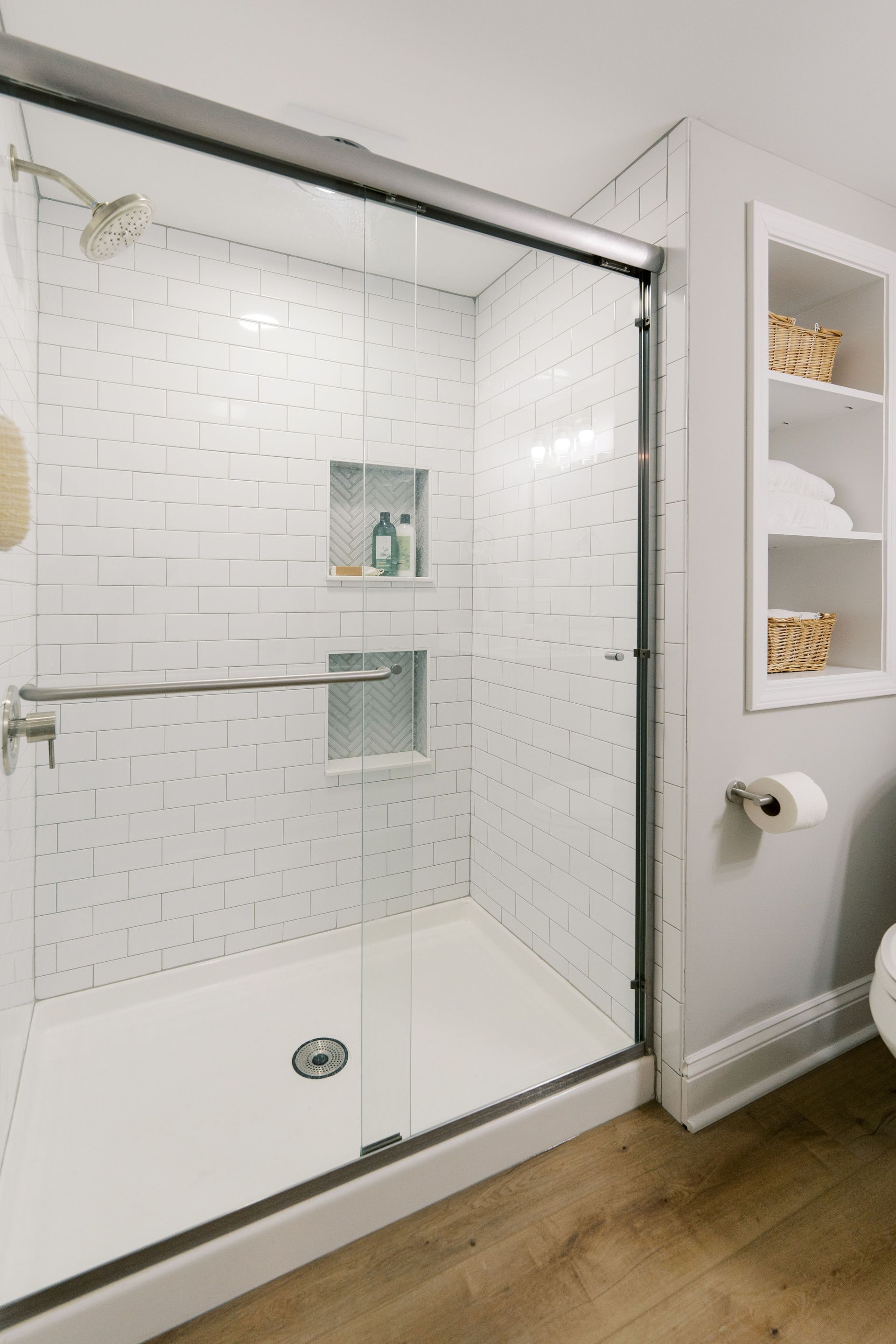 White tiled shower with glass sliding doors, built-in shelves on the side with baskets and towels.