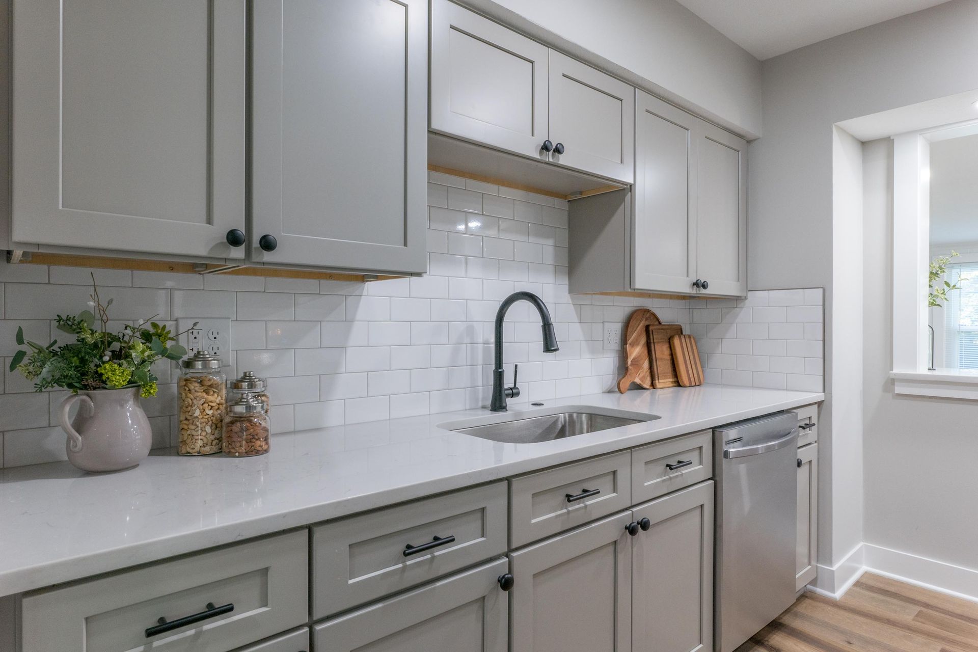 Gray kitchen cabinets with white countertops, a stainless steel sink, and black faucet.