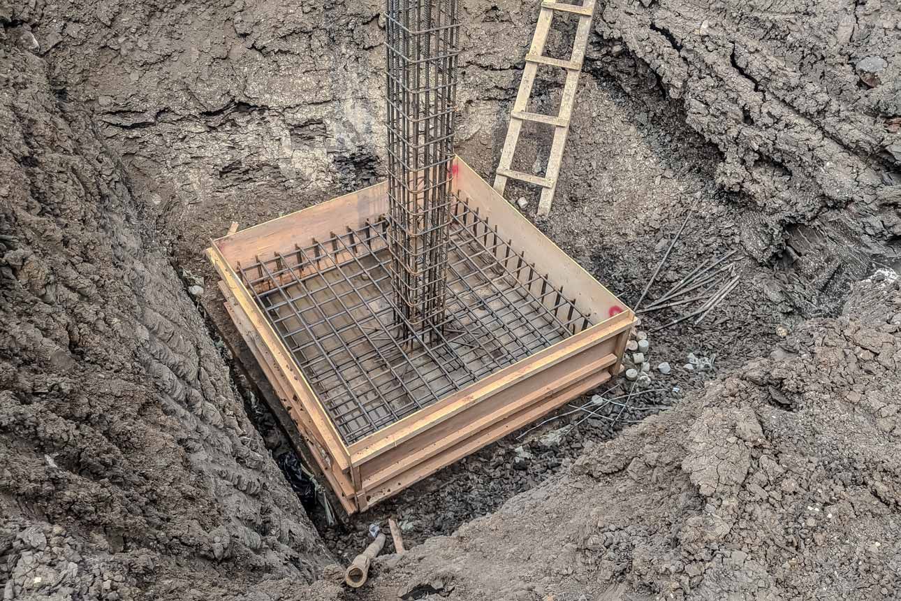 Wooden foundation form with steel rebar in a dug foundation pit, with a ladder leaning in nearby.