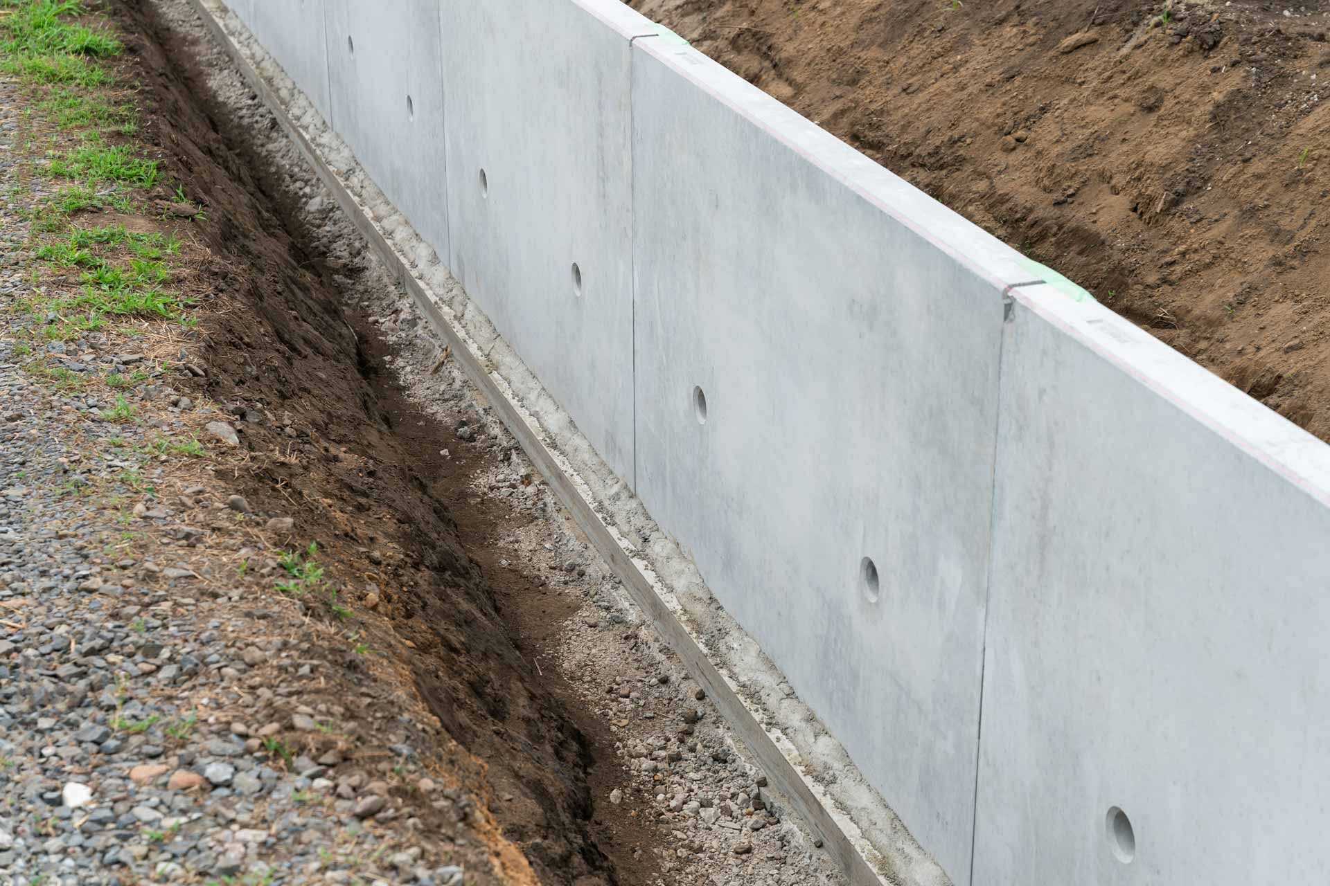 Concrete retaining wall beside a gravel path, with a dirt slope and erosion trench along the base