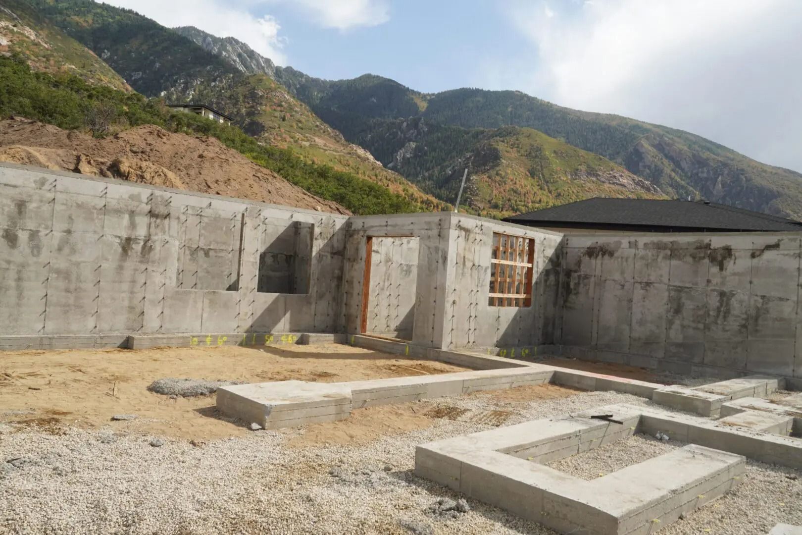 Concrete foundation site with retaining walls, gravel, and mountain backdrop under a cloudy sky