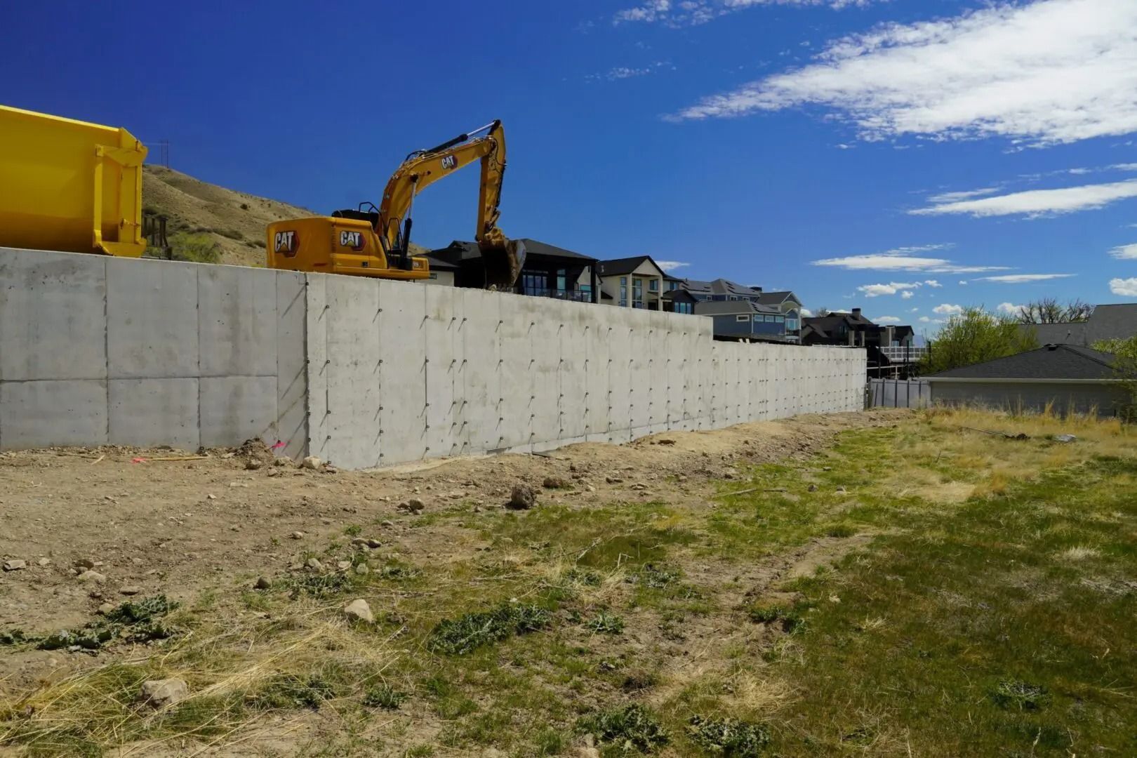 Construction site with excavator and yellow equipment behind a concrete retaining wall under a blue sky