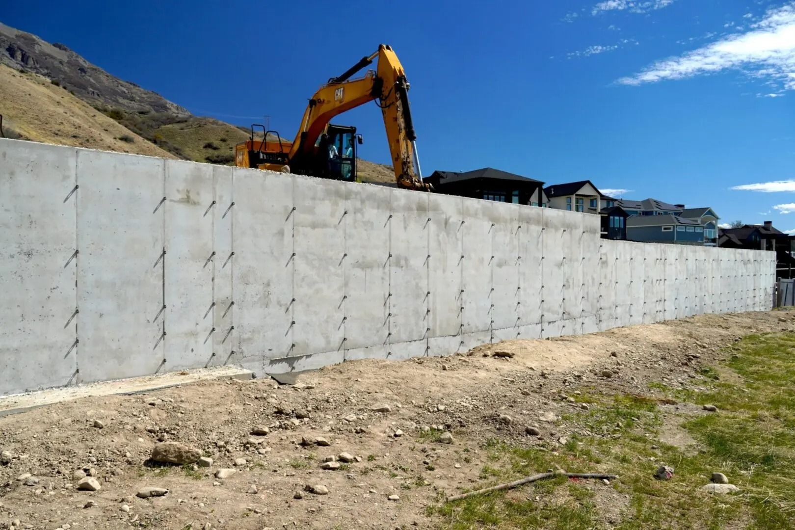 Excavator working behind a tall concrete retaining wall on a sunny hillside construction site.