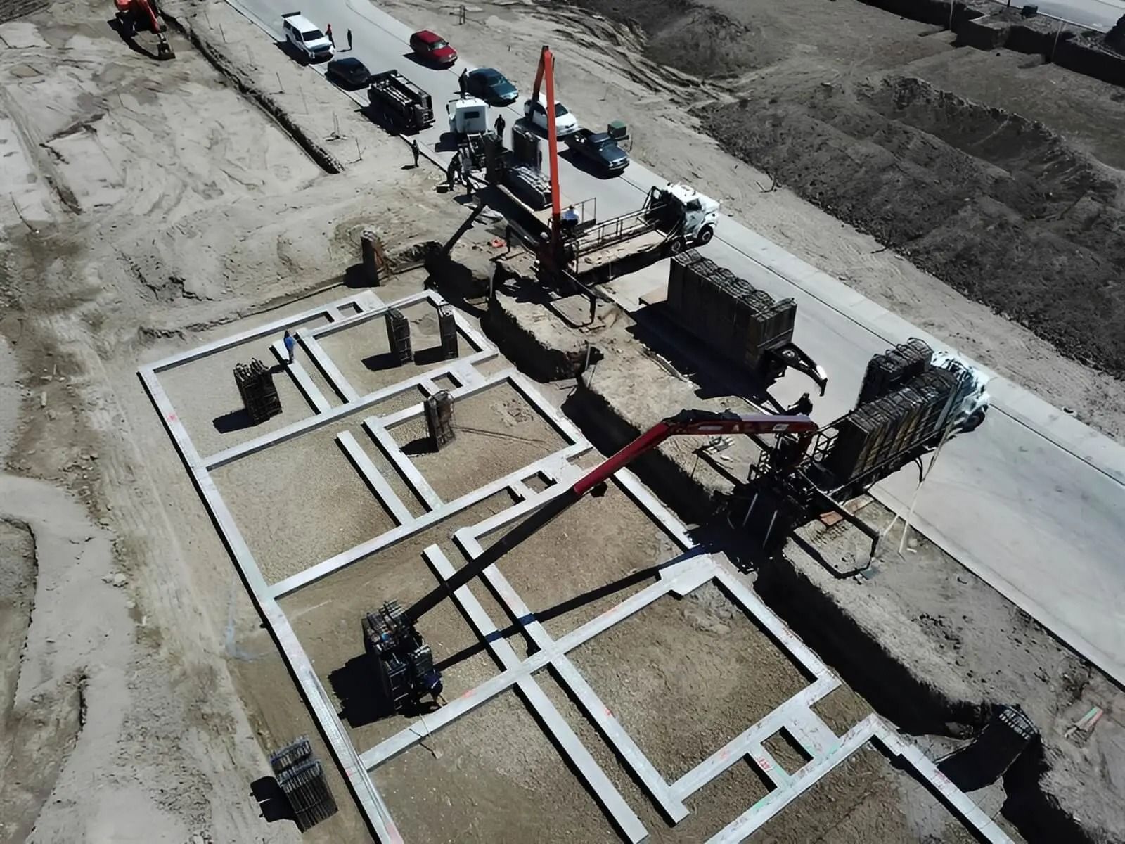 Aerial view of a construction site with foundation walls, heavy equipment, and workers near a paved road