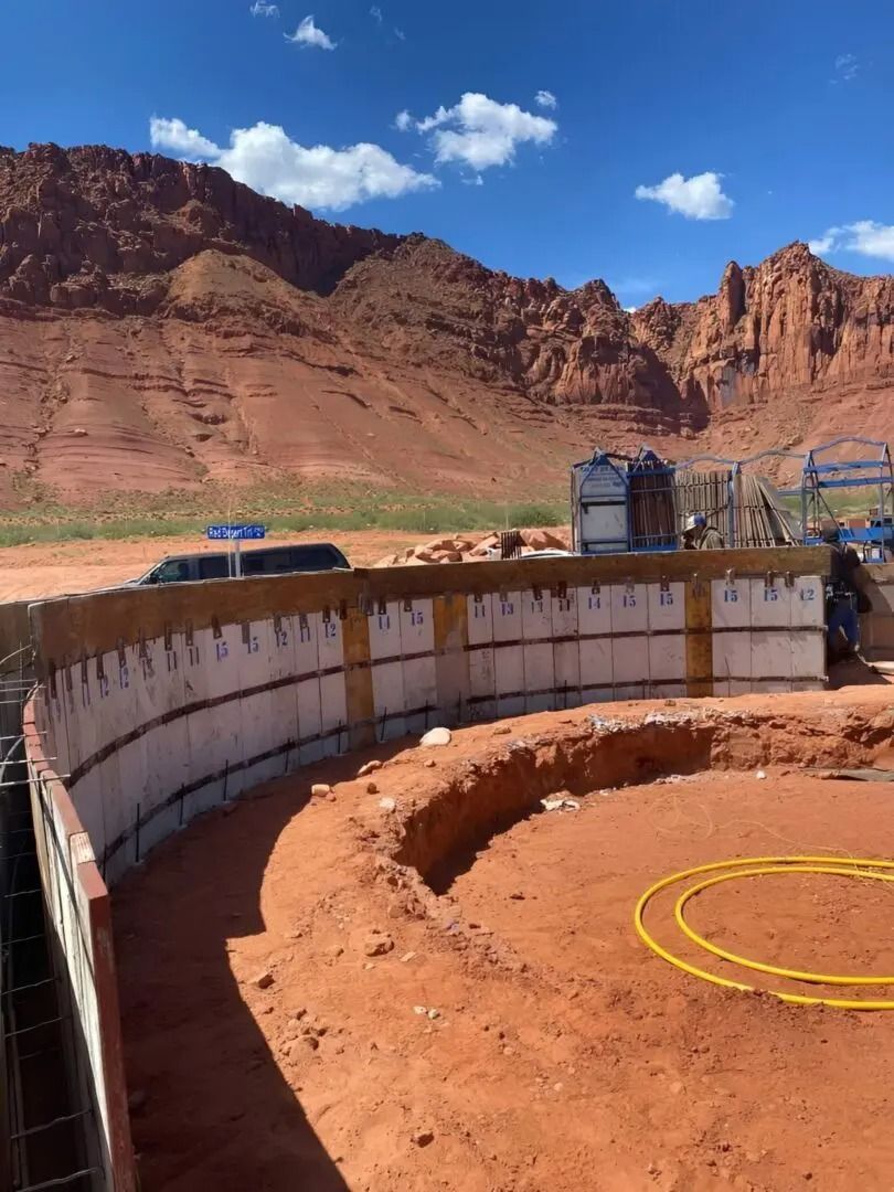 Curved concrete wall beside a red dirt site with yellow hose, backed by rocky cliffs and blue sky