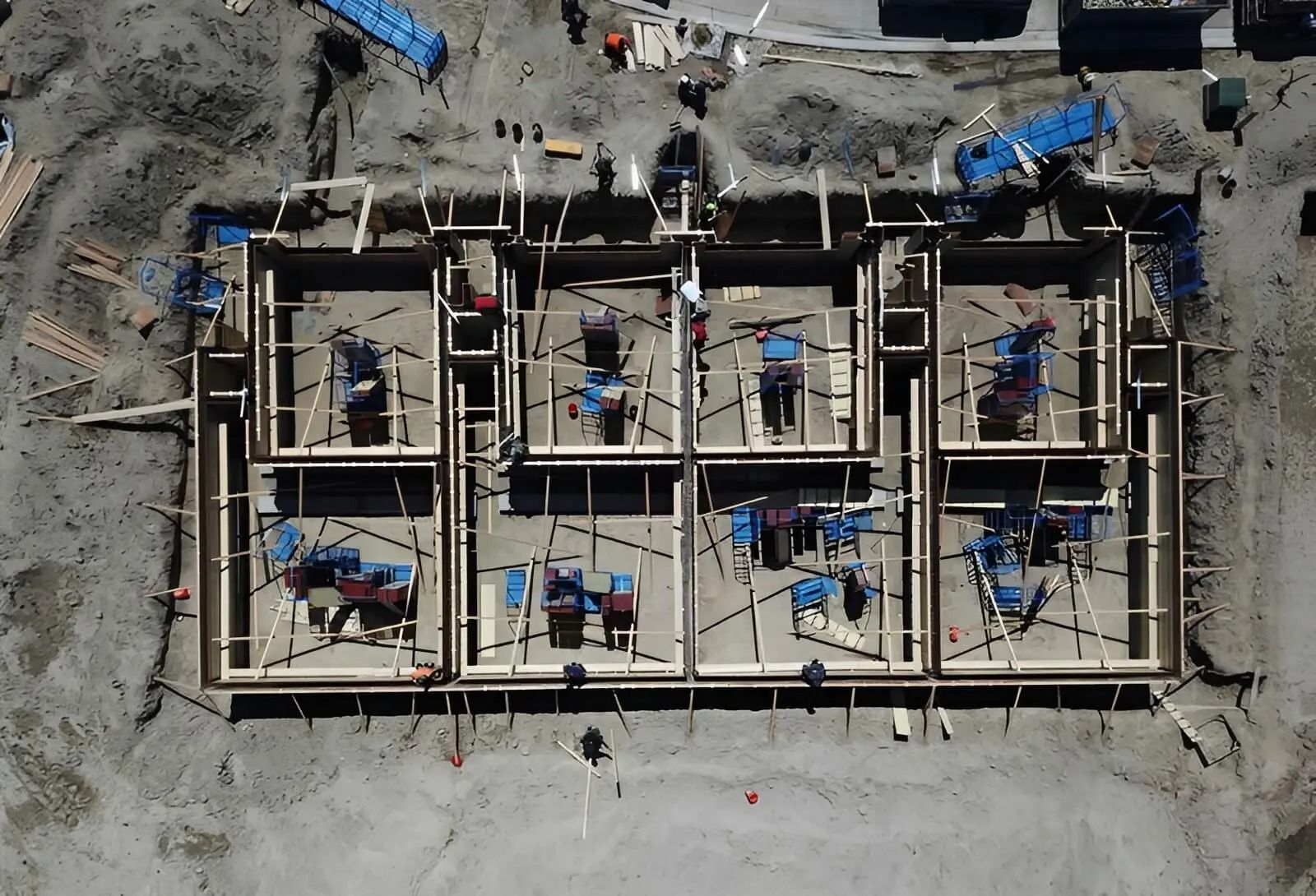 Overhead view of a construction site with workers, wooden framing, and blue machinery on concrete.