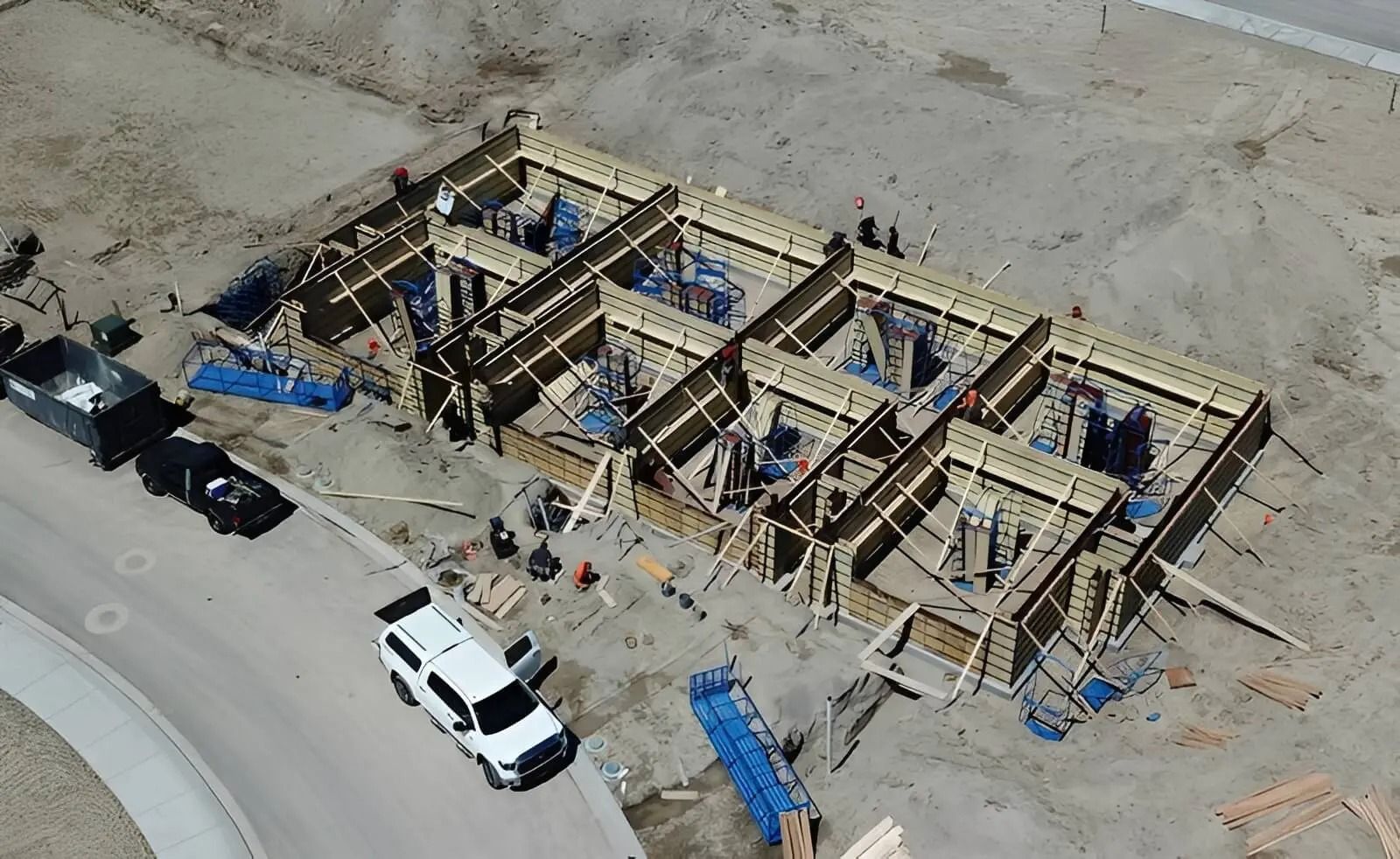 Aerial view of a wooden building frame under construction with workers, trucks, and blue tarps on a dirt site