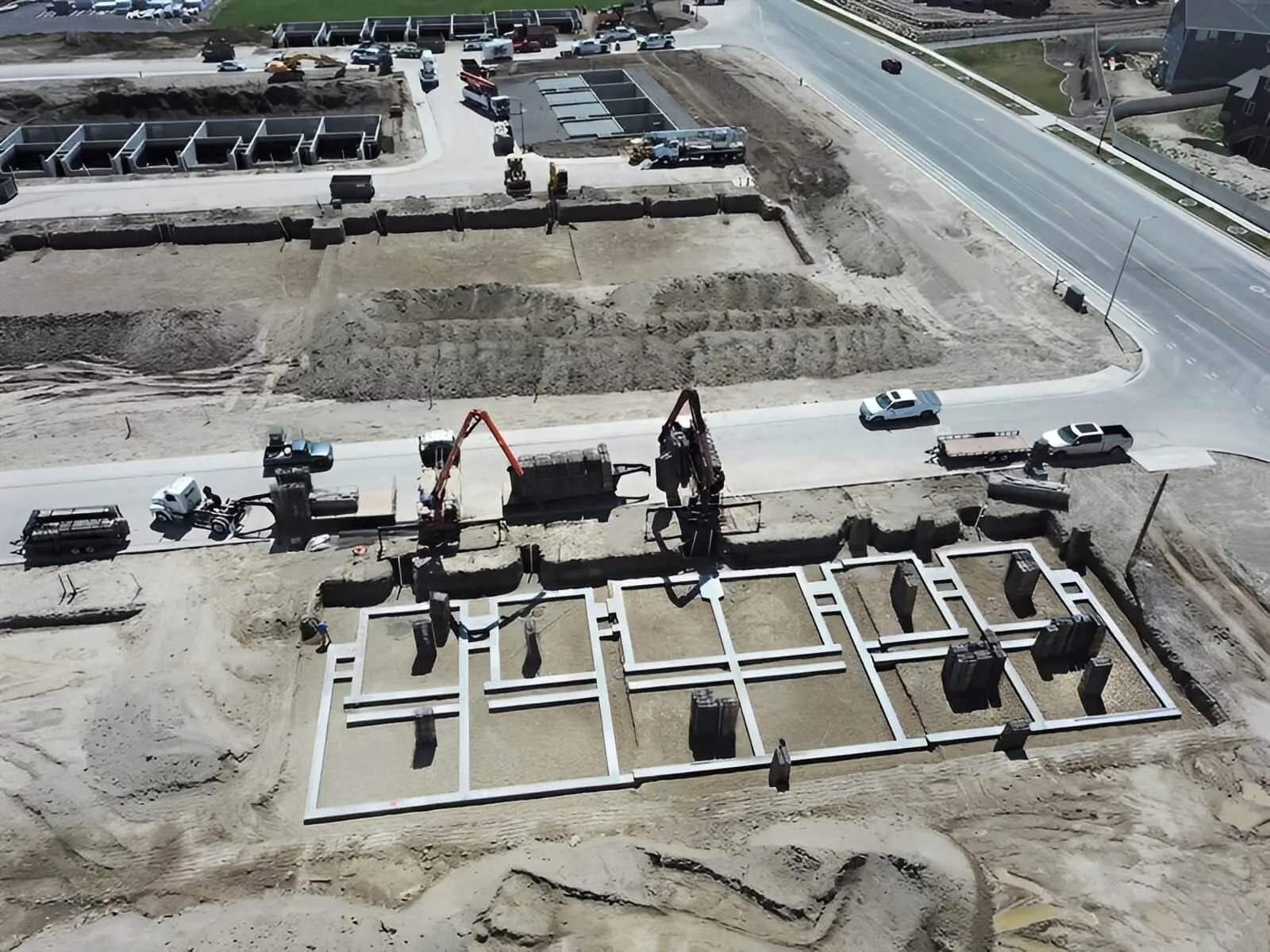 Construction site with foundation trenches, excavator, trucks, and workers beside a road