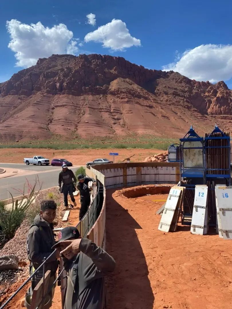 People near a curved fence in a red desert canyon with trucks and a blue structure under a cloudy sky