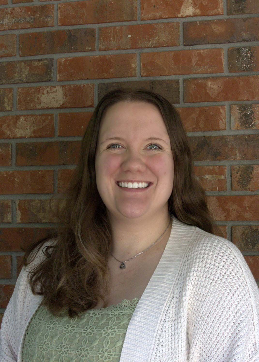 Smiling woman in a green top and white cardigan standing before a brick wall.