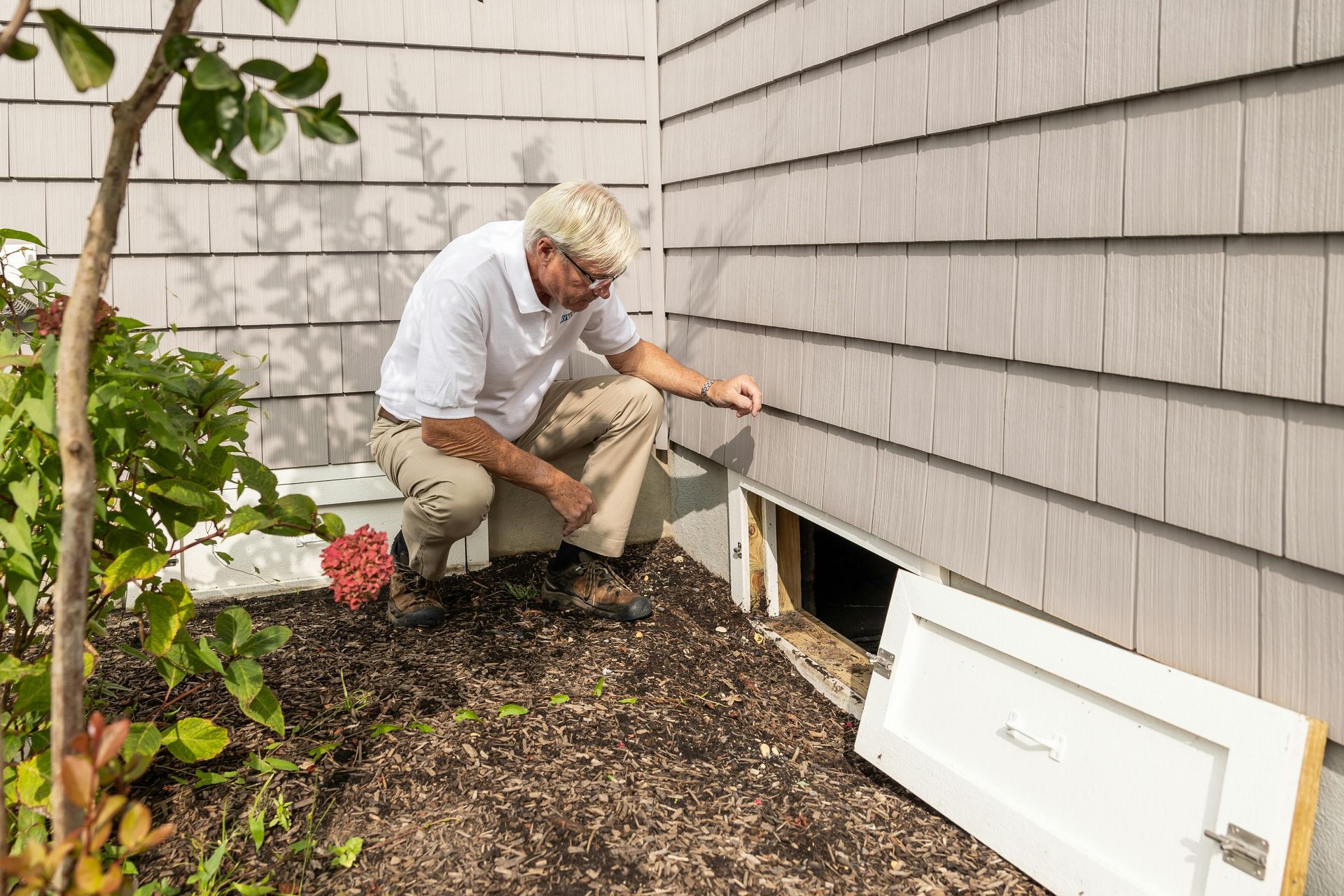Man inspecting crawl space door on the side of a house with gray siding. Brown pants, white shirt, and a brown yard.