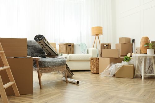 Living room filled with moving boxes, wrapped chair, and packed furniture on a wood floor
