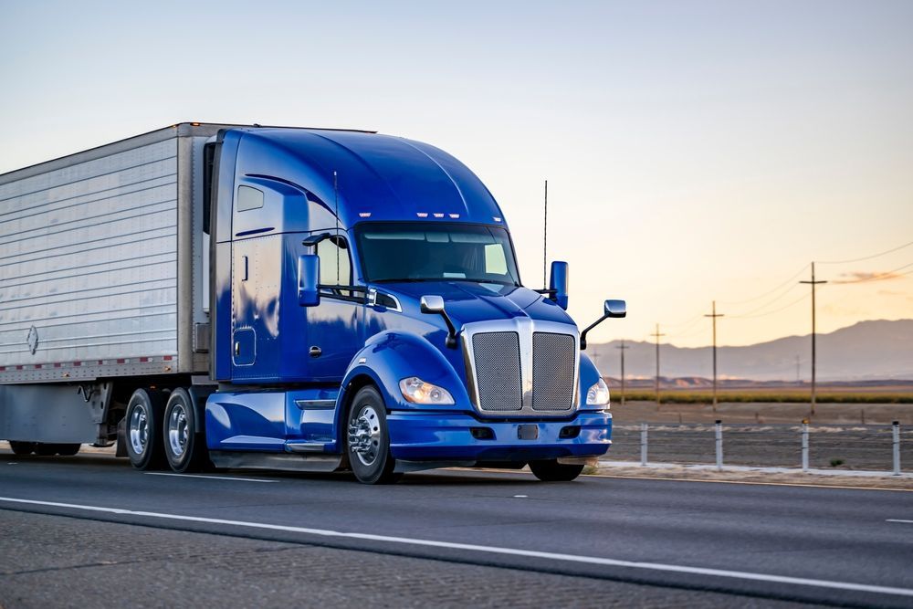 Blue semi-truck driving on a highway at sunset, hauling a silver trailer.