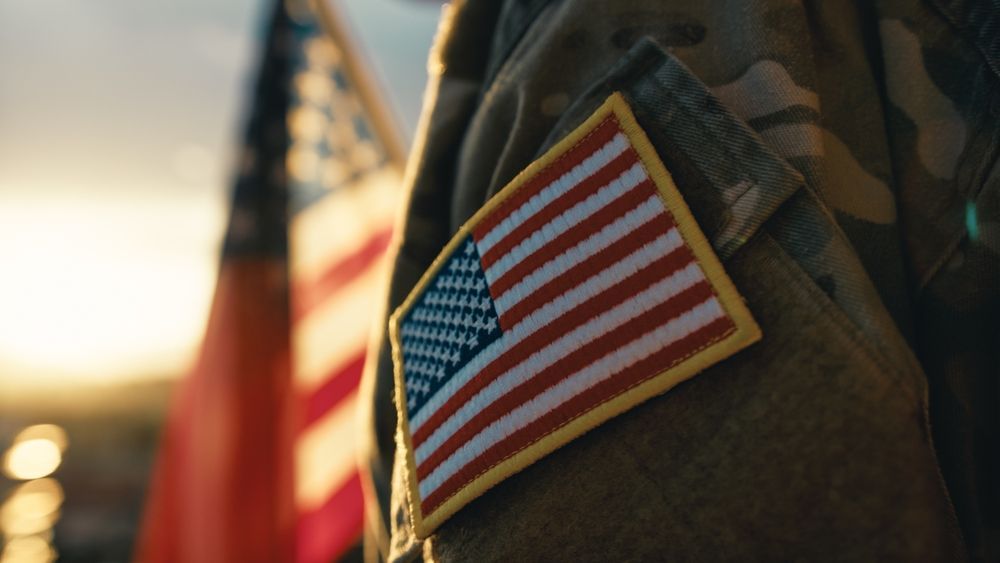American flag patch on a camouflage uniform at sunset, with a blurred flag in the background