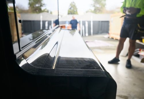Inside a garage, a glossy black piano sits in the foreground with two people blurred in the background.