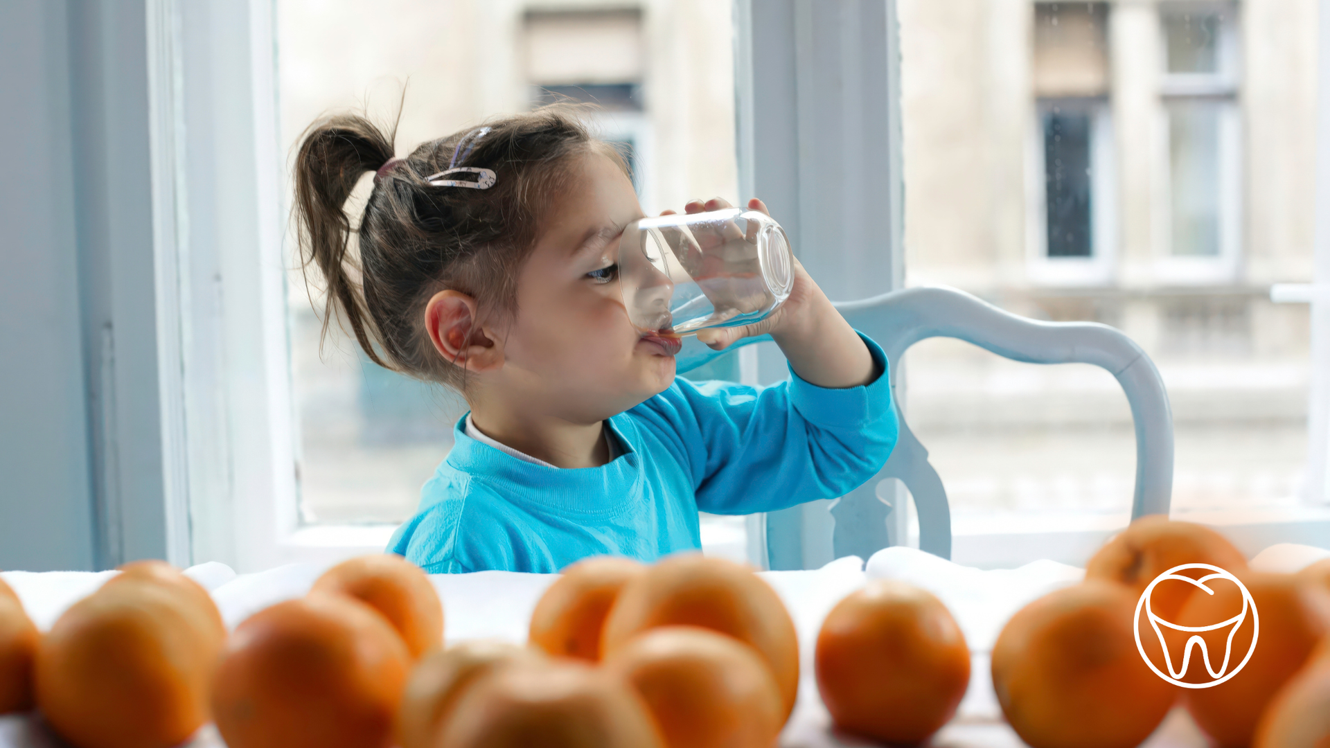 Young girl with a ponytail drinks water from a glass, oranges on the table in front of her.