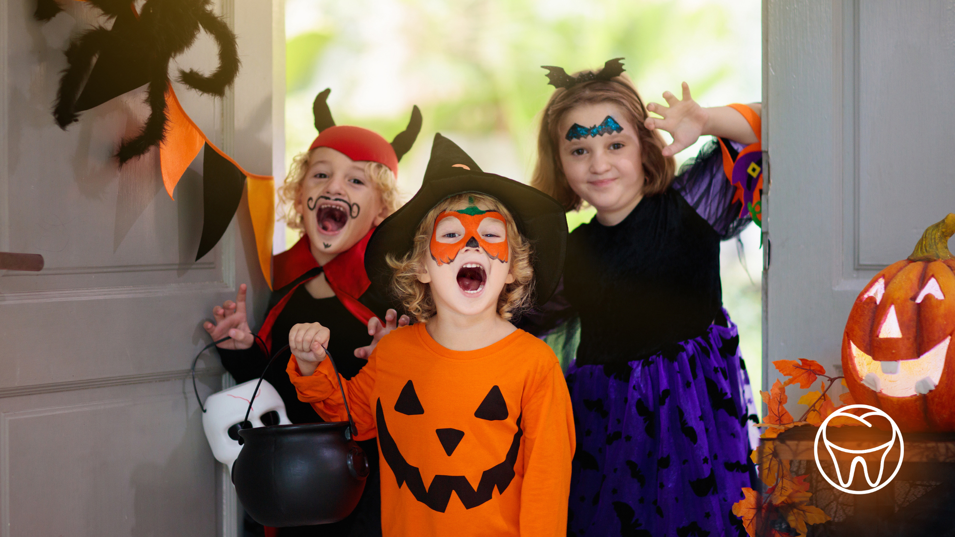 Three children in Halloween costumes excitedly stand at a door: witch, devil, and pumpkin.
