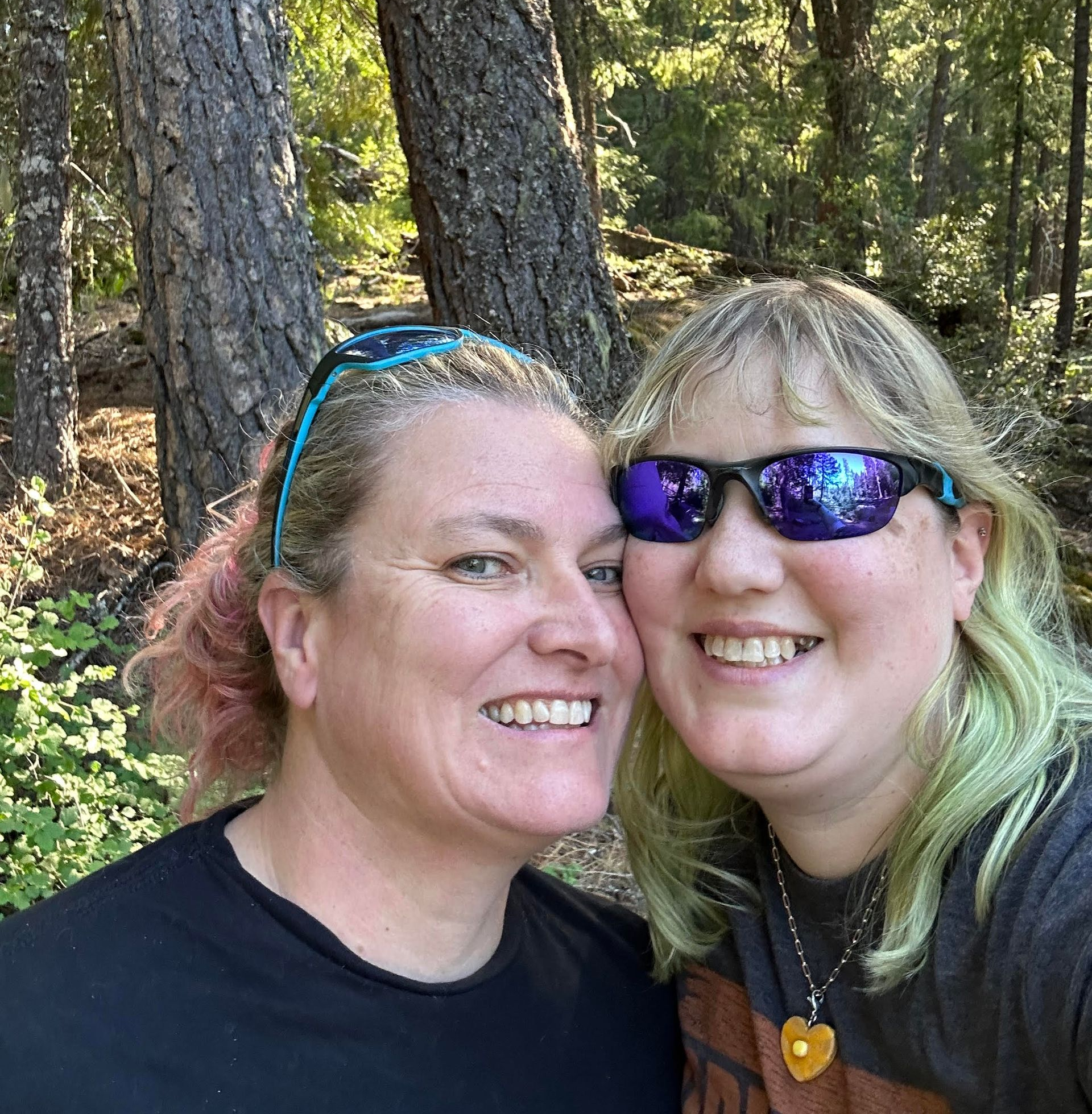 Two smiling women in a forest setting, one with pink hair, the other with green hair and sunglasses, close together.