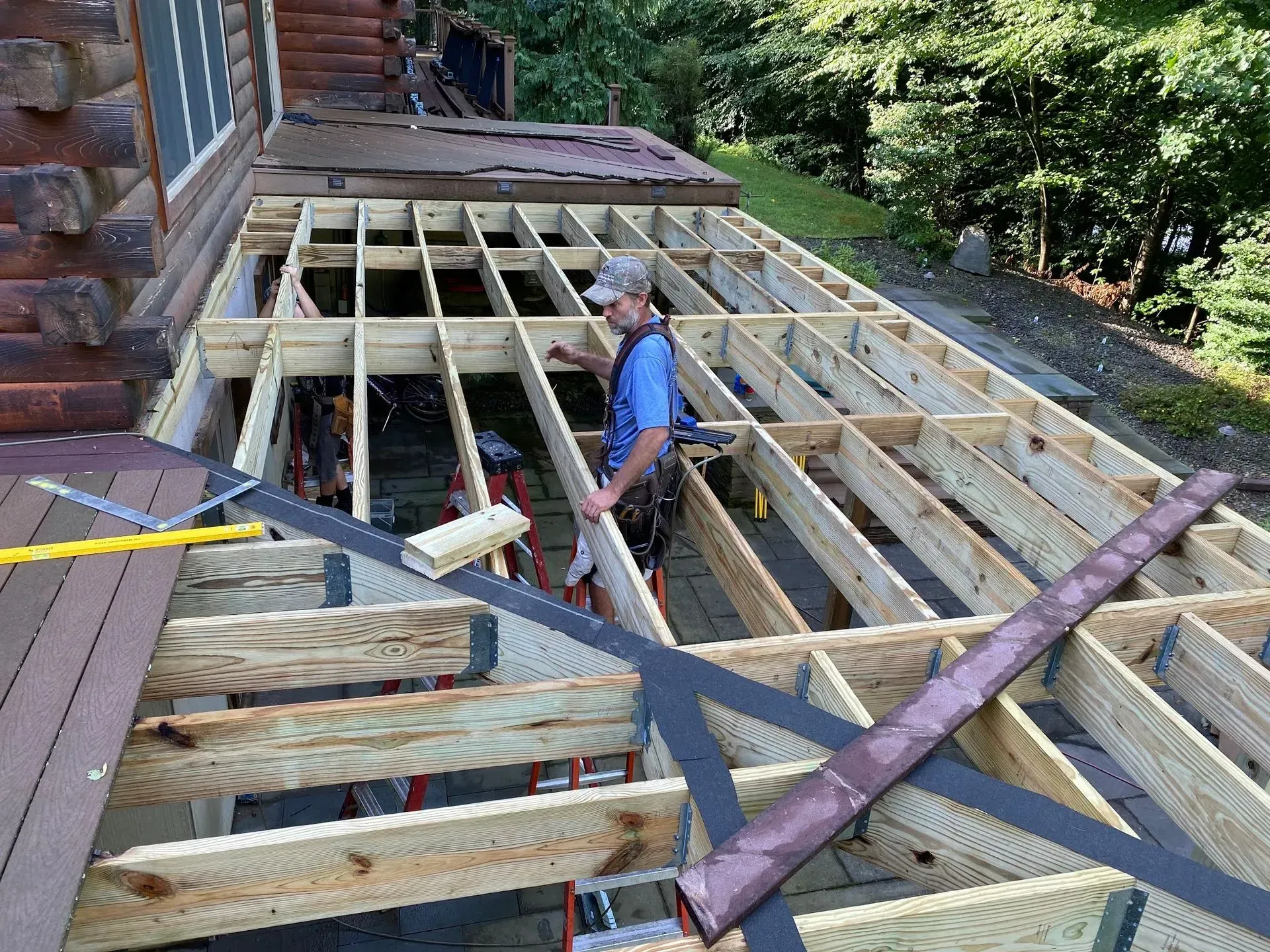 Worker framing a wooden deck beside a house, with exposed joists and construction materials.