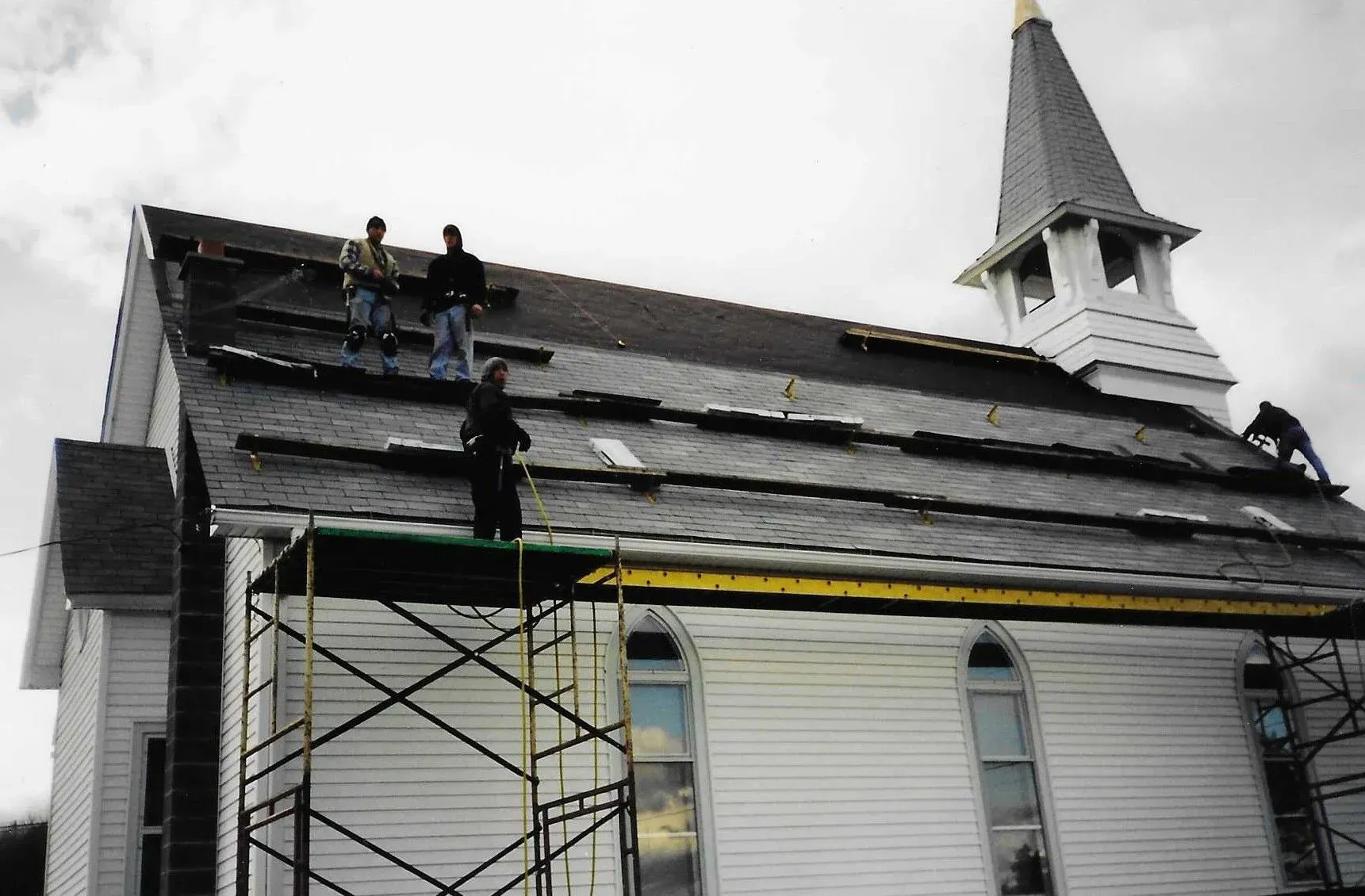 Workers repairing a church roof beside a steeple, with scaffolding and black shingles visible