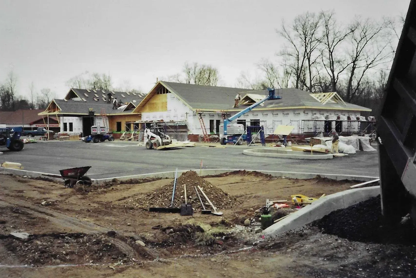 Construction site with a partially built house, dirt foreground, and workers near a row of buildings.