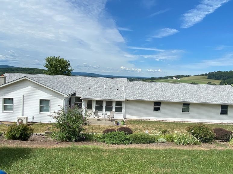White farmhouse with gray roof and garden in front, set against rolling green hills under a blue sky