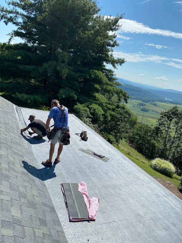 Person kneels on a sloped roof, working near tools with a scenic valley and trees in the background.