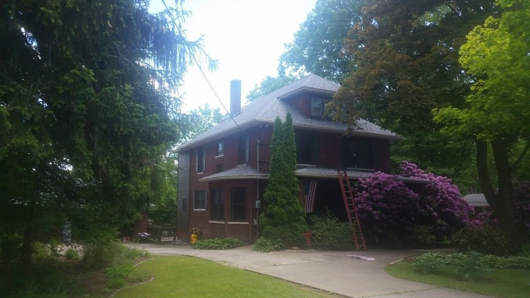 Two-story house with wraparound porch, surrounded by trees and flowers along a driveway