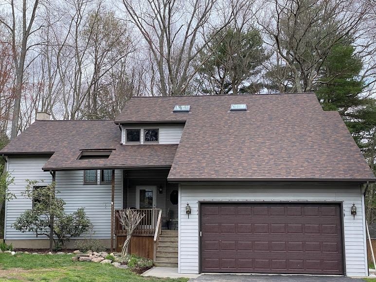 Suburban two-story house with brown roof, white siding, and a dark garage door surrounded by bare trees.