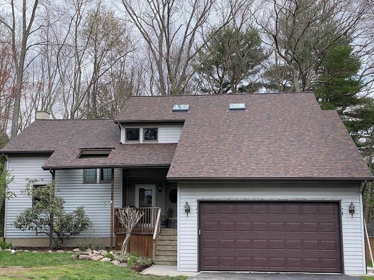 Suburban two-story house with brown roof, white siding, and a dark garage door surrounded by bare trees.