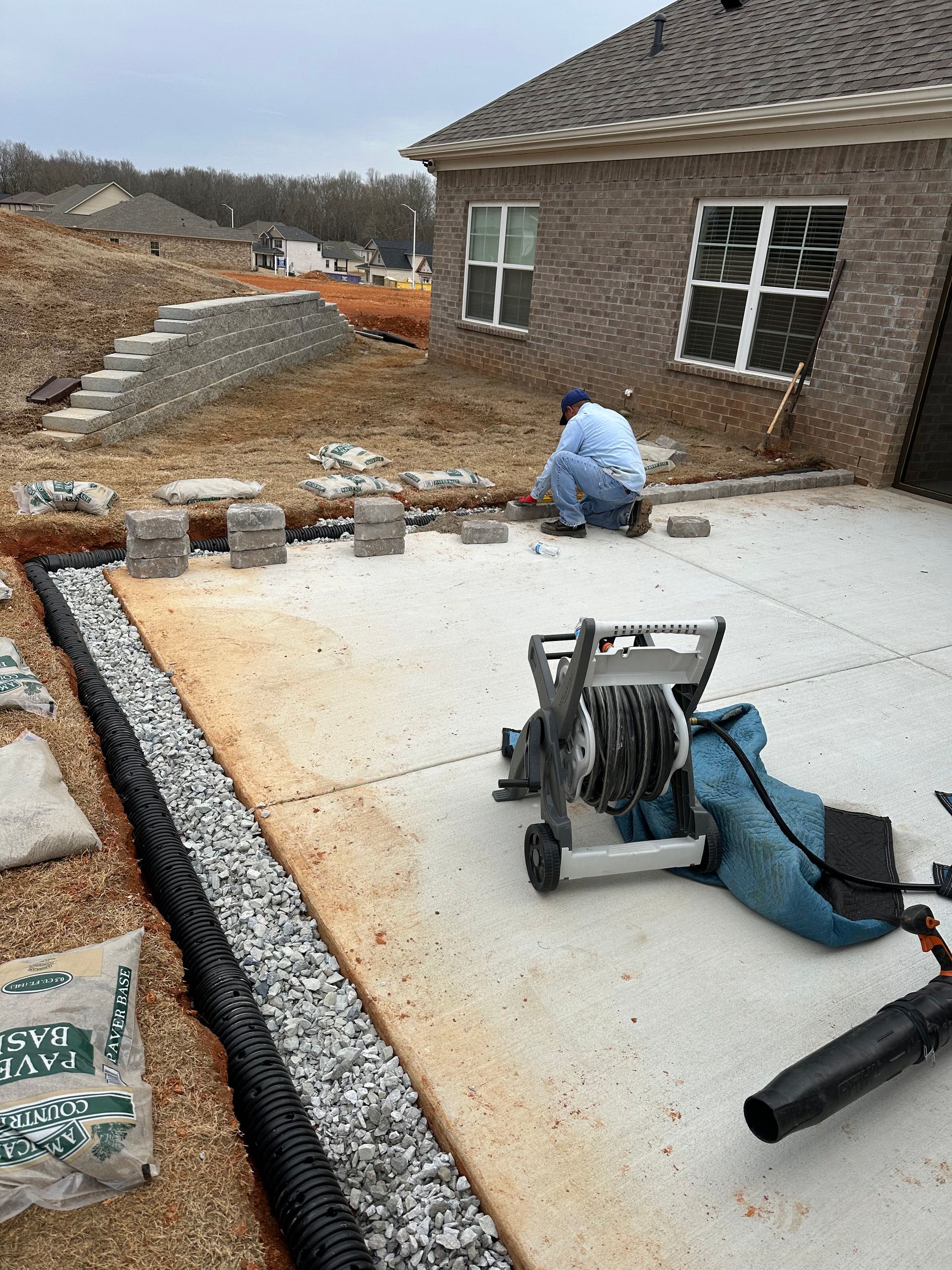 Man laying pavers next to a drainage trench on a patio near a brick house.