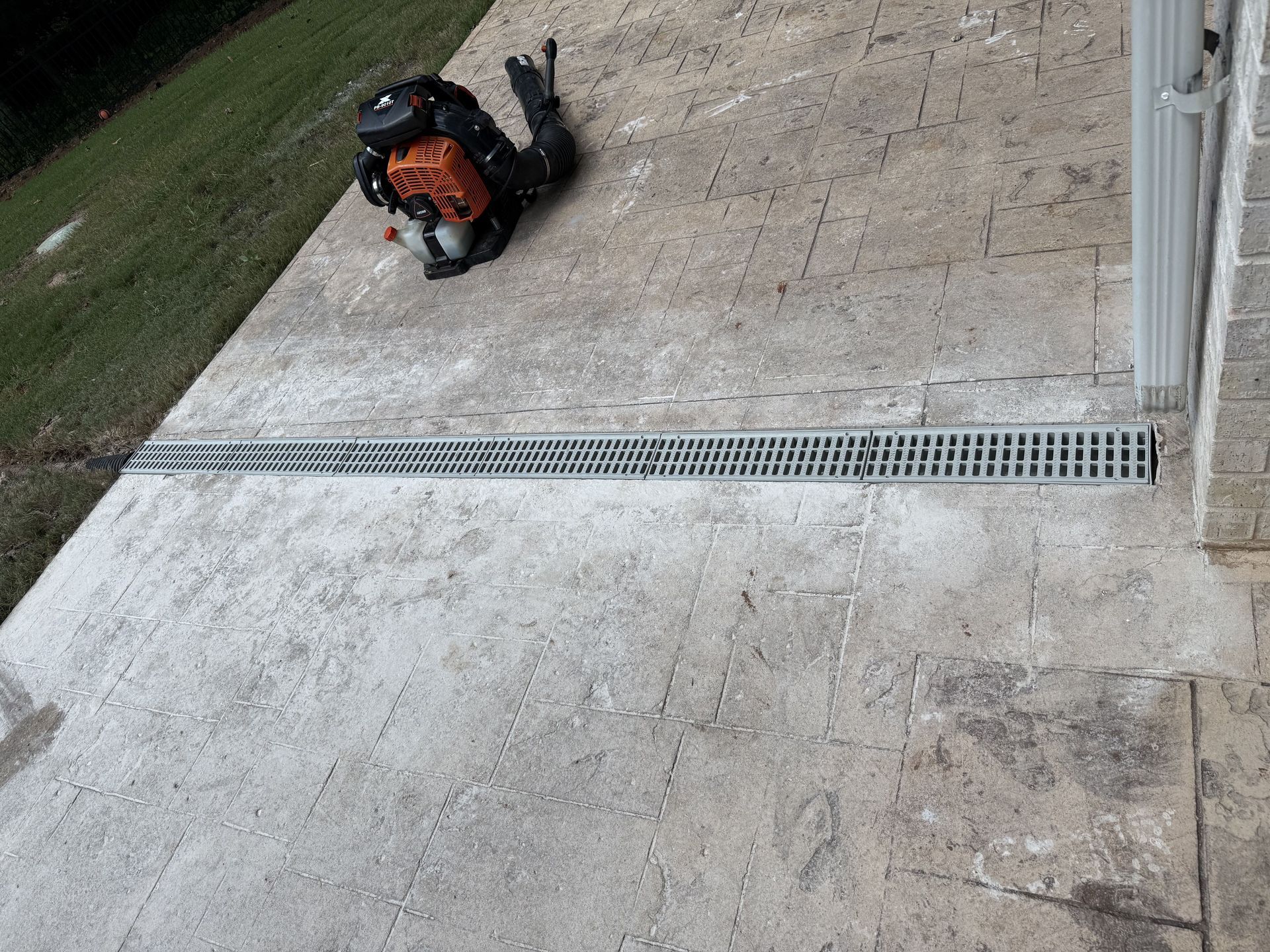 A concrete patio with a drainage grate, backpack, and building. Green grass to the left.