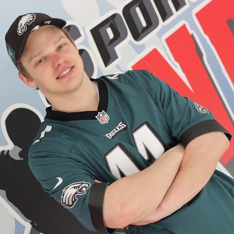 Man in Eagles jersey and hat, arms crossed, smiling in front of a sports-themed background.