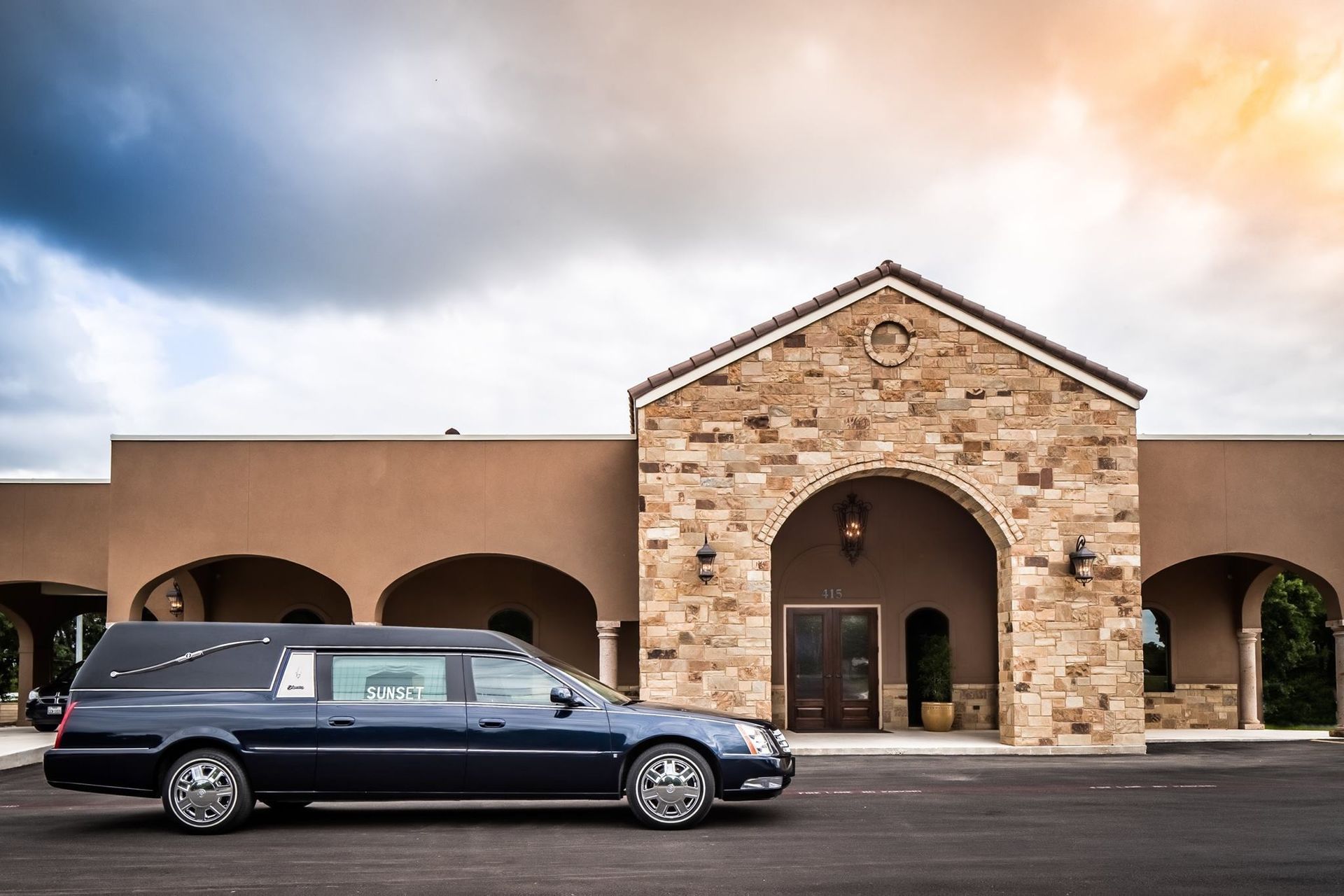 Dark blue hearse parked in front of a stone building with arches under a cloudy sky.